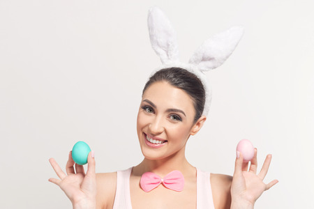 Studio shot of a smiling young woman wearing bunny ears and holding a of Easter eggs on light backgroundの写真素材