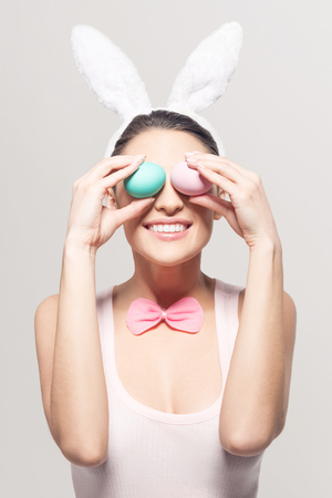 Studio shot of a happy young woman wearing bunny ears and holding up a colorful Easter egg in front of her eyeの写真素材