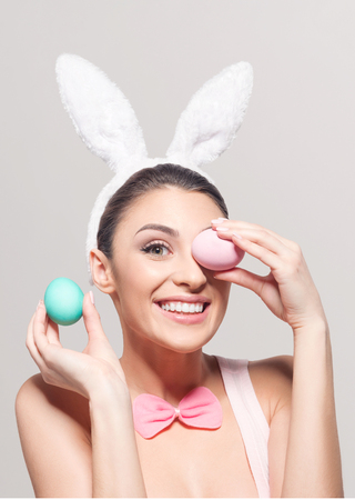 Studio shot of a smiling young woman wearing bunny ears and holding a of Easter eggs on light backgroundの写真素材