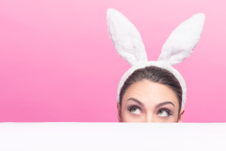 Studio shot of a young woman wearing bunny ears and peeking  up away on copy space over a surfaceの写真素材