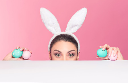 Studio shot of a young woman wearing bunny ears peeking over a surface and holding easter eggsの写真素材