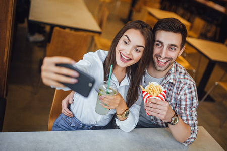A young lovers couple makes selfi on a mobile phone with mojito and a hamburger and smiles in a fast food cafe.の写真素材