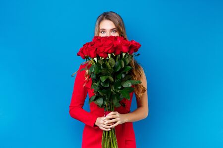 cute girl covering face with bouquet of red roses on blue background.の写真素材
