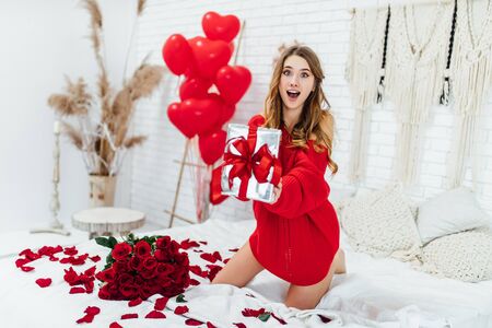 emotional girl showing present box and looking at the camera while sitting on the bed with st valentines gifts.の写真素材