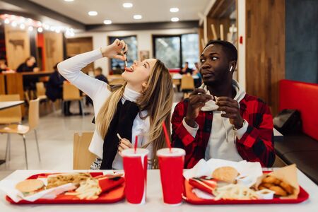 Two young students having fun during lunch at bar, sitting at table with drinks and fast foodの写真素材