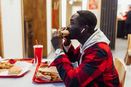 African american man bites hamburger, handsome and young afro man in a stylish shirt holding a burger on a fast fooの写真素材