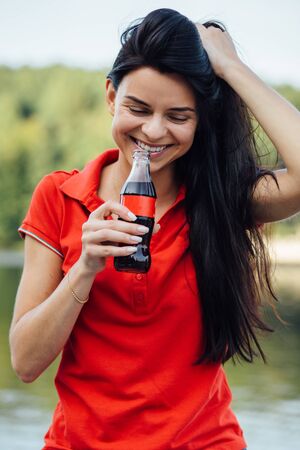 Close up of beautiful young smiling woman in red t-shirt enjoying drinking colaの写真素材