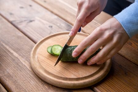 hands of a girl cut cucumber on a wooden table, the process of making vegetarian salad, close-up cutting of vegetables and greensの写真素材