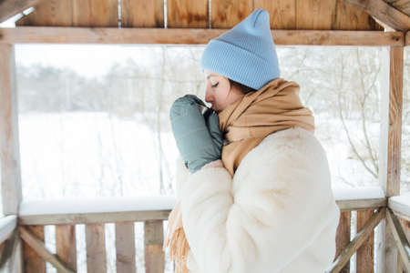 beautiful girl in a white fur coat drinks warm tea in winter in a snowy forestの写真素材