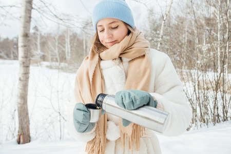 beautiful girl in a white fur coat pours warm tea in winter in a snowy forestの写真素材