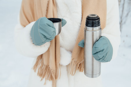 A girl in warm mittens holding a steel bottle with tea in a snowy forest in winter