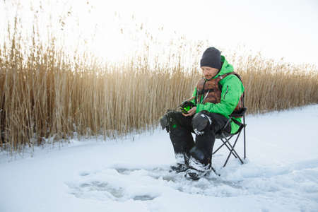 a young man on a winter fisherman sits on a chair and fishes and a fishing rodの写真素材