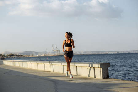 beautiful fitness girl in shorts and sports top runs on the waterfront by the sea during sunsetの写真素材