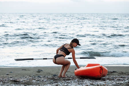 beautiful sports girl in a black swimsuit goes with an orange kayak to swim in the sea at sunsetの写真素材