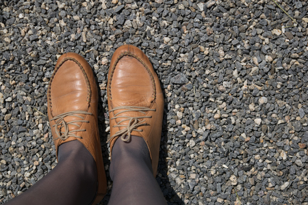 Female feet in brown leather shoes and kapron pantyhose of mocha color on a background of fine gravelの写真素材