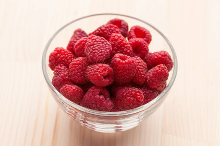 Raspberries in a glass bowl on wooden table, close-up. 