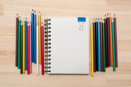 Checked blank notebook with blue binder clip and multicolored pencils on wood background.の写真素材