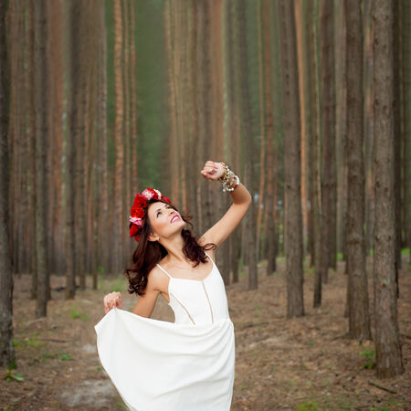 Fairytale in the forest. Beautiful  woman wearing white dress and with red roses hairband in pine forest.の写真素材