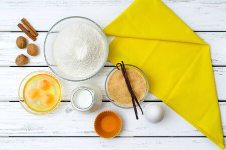 Baking in rural kitchen - recipe ingredients (eggs, flour, milk, sugar, vanilla, walnuts, cinnamon) with yellow kitchen napkin on white wooden table from above.の写真素材
