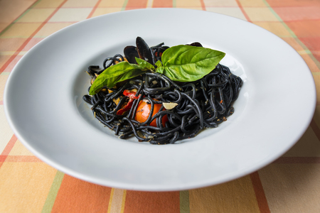 Italian black spaghetti with cuttlefish ink, mussels, tomatoes and green basil on the table with orange plaid tablecloth at sunlight.の写真素材