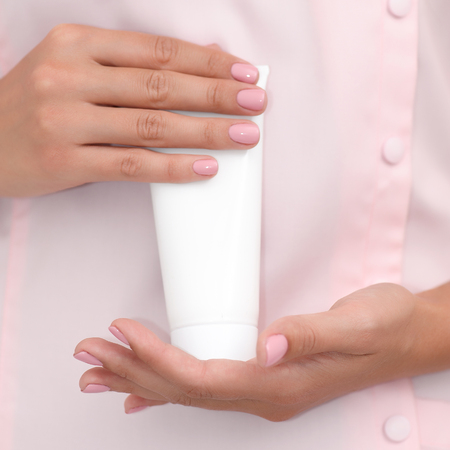Hand skin care. Female hands holding one white cream tube against body in pink cloth, beautiful beautician woman hands with manicured nails close-up background, shallow DOF, square composition.の写真素材