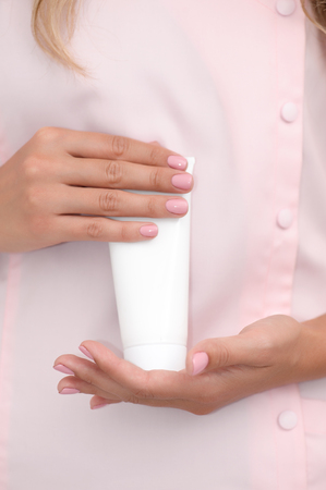 Hand skin care. Female hands holding one white cream tube against body in pink cloth, beautiful beautician woman hands with manicured nails close-up background, shallow DOF, soft focus.の写真素材