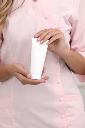 Hand skin care. Female hands holding one white cream tube against body in pink cloth, beautiful beautician woman hands with manicured nails close-up background, shallow DOF, soft focus.の写真素材