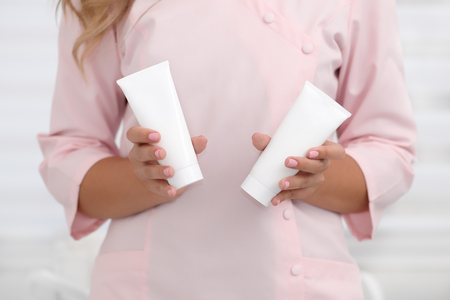 Hand skin care. Female hands holding two white cream tubes against body in pink cloth, beautiful beautician woman hands with manicured nails close-up background, shallow DOF, soft focus.の写真素材