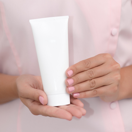 Hand skin care. Female hands holding one white cream tube against body in pink cloth, beautiful beautician woman hands with manicured nails close-up background, shallow DOF, square composition.の写真素材