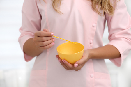 The hands of a beautician stir the ingredients in a yellow cup for applying the mask on the skin. Cosmetology concept. Horizontal composition. Shallow dof, soft focus.の写真素材