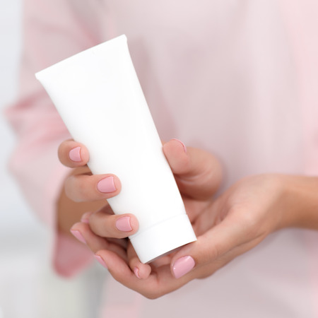 Hand skin care. Female hands holding one white cream tube against body in pink cloth, beautiful beautician woman hands with manicured nails close-up background, shallow DOF, square composition.の写真素材