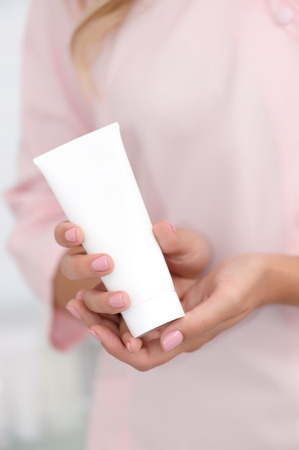 Hand skin care. Female hands holding one white cream tube against body in pink cloth, beautiful beautician woman hands with manicured nails close-up background, shallow DOF, soft focus.の写真素材