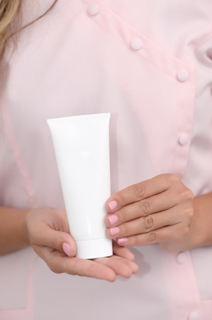 Hand skin care. Female hands holding one white cream tube against body in pink cloth, beautiful beautician woman hands with manicured nails close-up background, shallow DOF, soft focus.の写真素材