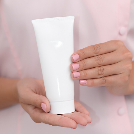 Hand skin care. Female hands holding one white cream tube against body in pink cloth, beautiful beautician woman hands with manicured nails close-up background, shallow DOF, square composition.の写真素材