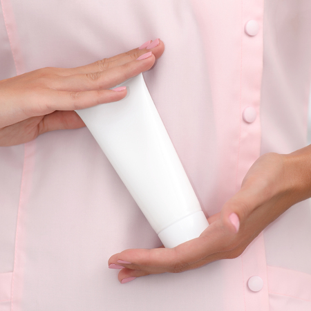 Hand skin care. Female hands holding one white cream tube against body in pink cloth, beautiful beautician woman hands with manicured nails close-up background, shallow DOF, square composition.の写真素材