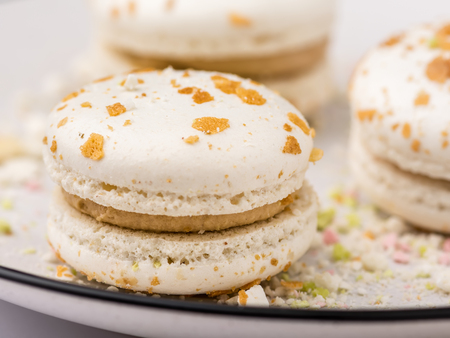 Three beige white macaroon cookies with multicolored crumb on white plate with blue border. Food background, shallow DOF, soft focus.の写真素材
