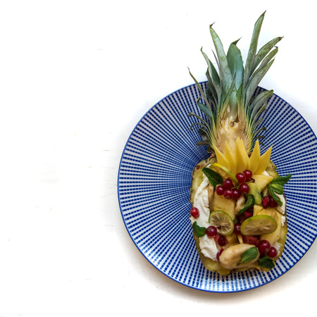 Pineapple decorated with exotic fruits. Close-up of fruit assortment at blue plate over white background. Mango, kiwi, pineapple, banana, lime, spearmint and red currant.の写真素材