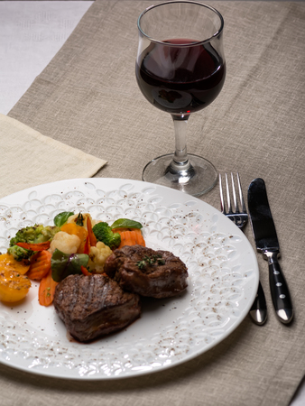 Grilled meat with vegetables in white plate, and red wine on the table. Restaurant table setting close-up.の写真素材