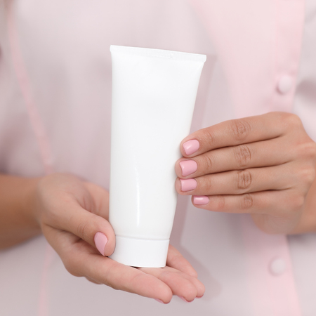 Hand skin care. Female hands holding one white cream tube against body in pink cloth, beautiful beautician woman hands with manicured nails close-up background, shallow DOF, square composition.の写真素材
