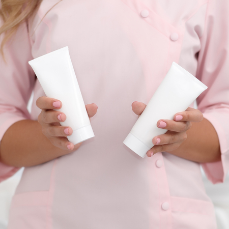 Hand skin care. Female hands holding two white cream tubes against body in pink cloth, beautiful beautician woman hands with manicured nails close-up background, shallow DOF, square composition.の写真素材