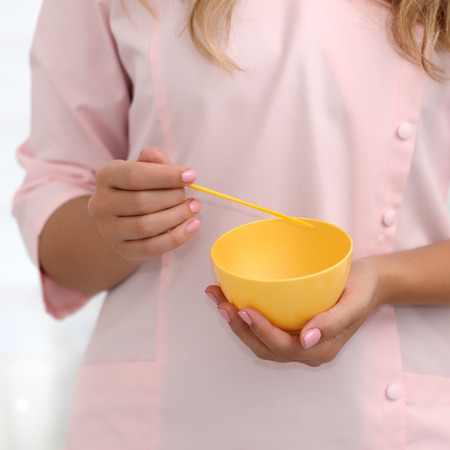 The hands of a beautician stir the ingredients in a yellow cup for applying the mask on the skin. Cosmetology concept. Square composition. Shallow dof, soft focus.の写真素材