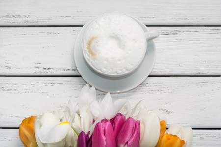 Bouquet of multicolored tulips in paper wrapper and cappuccino coffee with mil froth on white wooden table.の写真素材