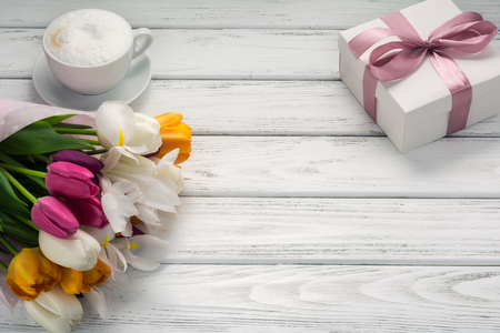 Bouquet of multicolored tulips in paper wrapper, gift box, cappuccino coffee with milk froth on white wooden table. Copy space. Beautiful breakfast on Mothers or Woman's day.の写真素材