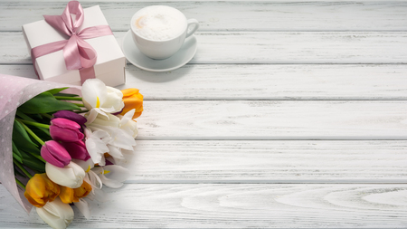 Bouquet of multicolored tulips in paper wrapper, gift box, cappuccino coffee with milk froth on white wooden table. Copy space. Beautiful breakfast on Mothers or Woman's day.の写真素材