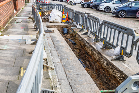 Open trench road construction with safety barriers and parked cars along urban streetの写真素材