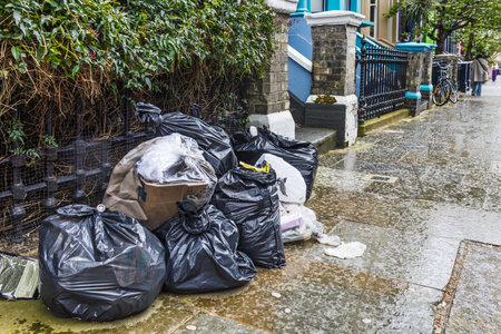 Overflowing Garbage Bags Left on Wet Sidewalk Next to Urban Streetの写真素材
