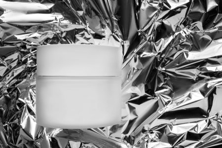Blank white cosmetic product jar against crumpled silver foil background