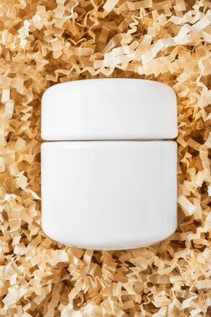 Blank white beauty product jar lying in yellow shredded packaging paper