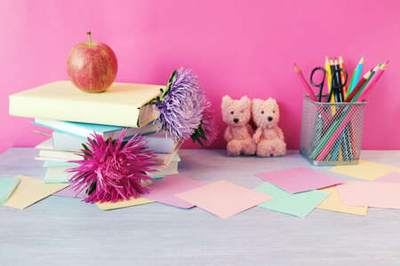 Stack of books with bookmarks of fresh flowers, apple, teddy bears, pencils and paper on a wooden...