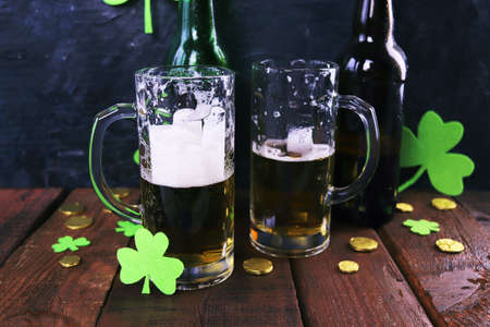 Patrick day, foamy beer in glass mugs and a bottle, gold coins on a wooden table, green shamrock on a dark background, party, congratulation, postcardの写真素材